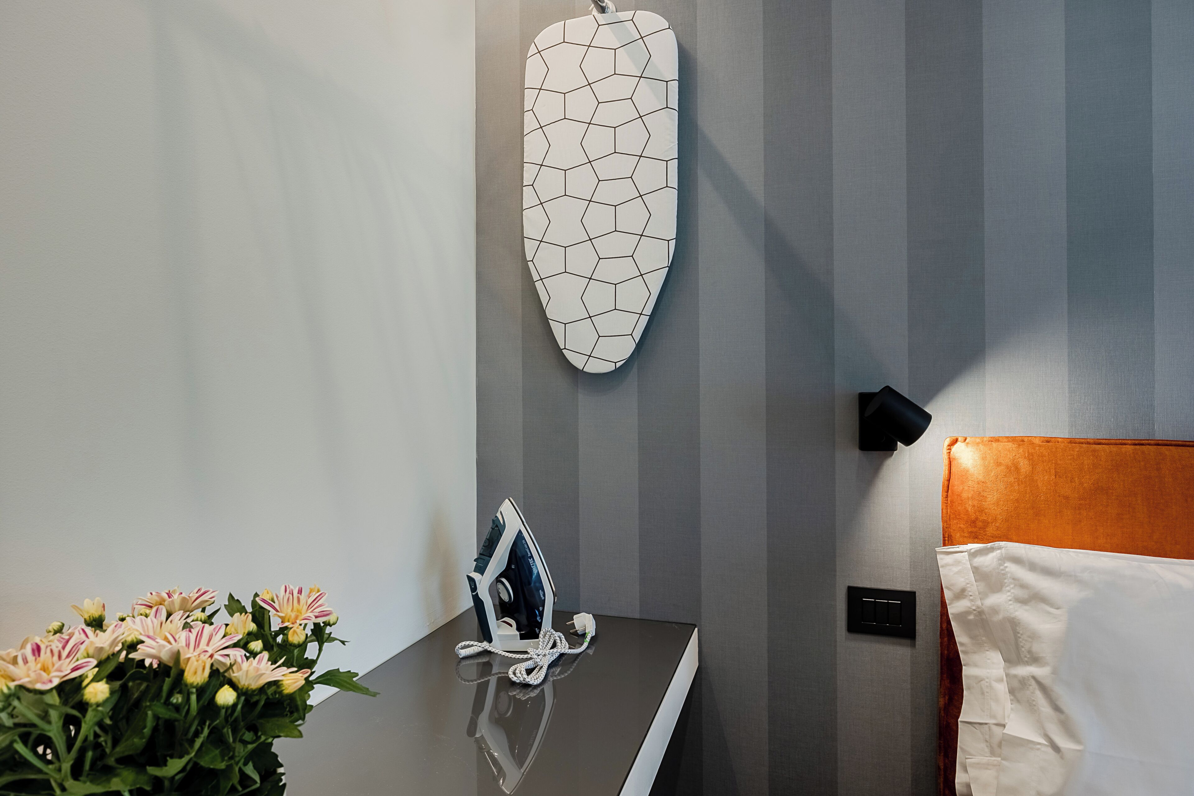 A hotel room with a bed, a bedside table with an iron, a wall-mounted heater, and a vase of flowers.