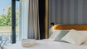 A hotel room with a bed, pillows, a nightstand, a window with curtains, and a view of buildings and greenery outside. - Il Pumo - Apulian Rooms Bari Moro 61 (Bari)