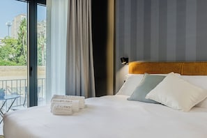 A hotel room with a bed, pillows, a nightstand, a window with curtains, and a view of buildings and greenery outside.
