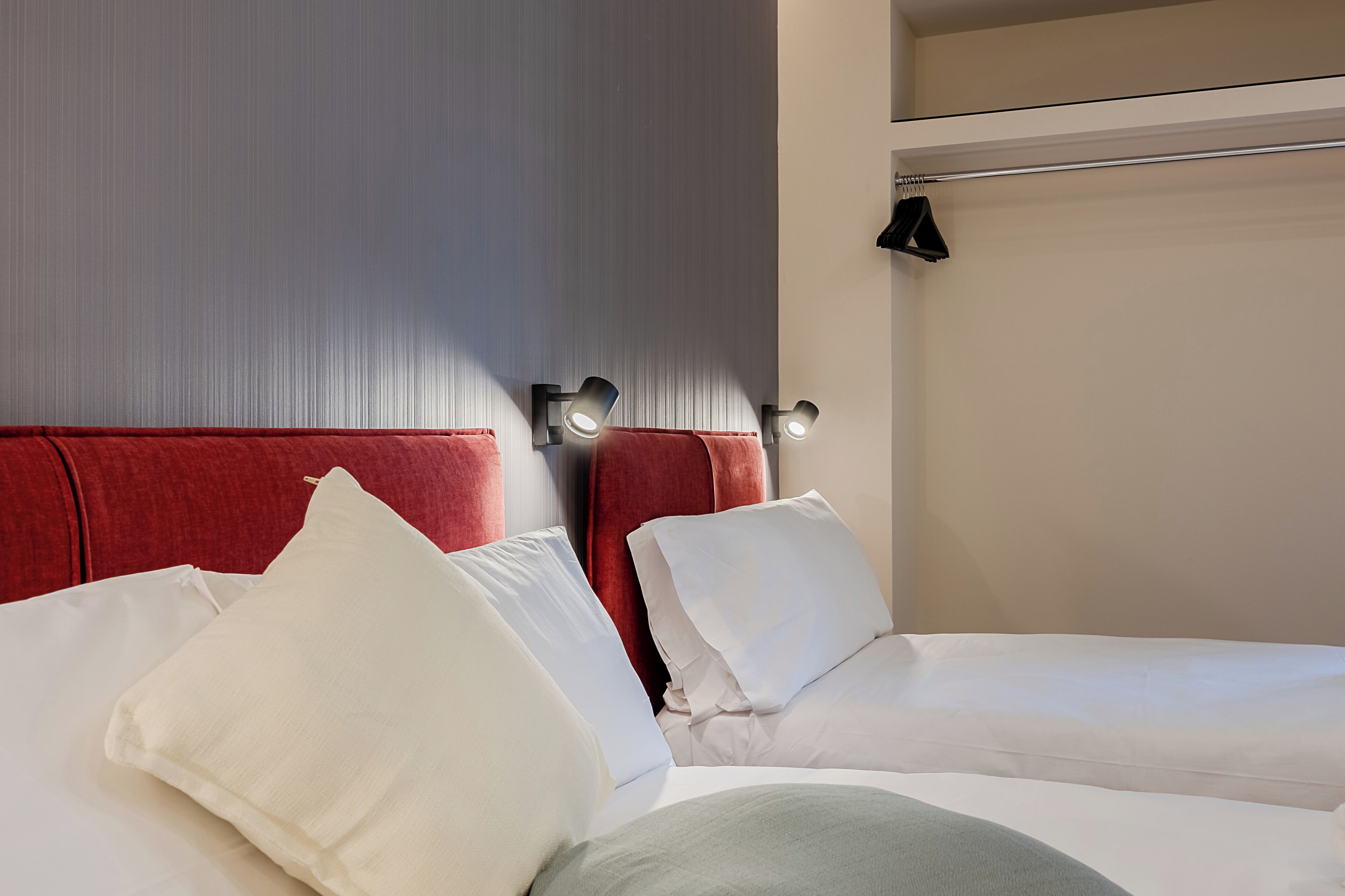A hotel room with two beds, each with white bedding and red headboards. There are wall-mounted lights above each bed.