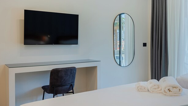 A hotel room with a flat-screen TV mounted above a desk, a round mirror on the wall, a chair, and a bed with rolled towels. - Il Pumo - Apulian Rooms Bari Moro 61 (Bari)