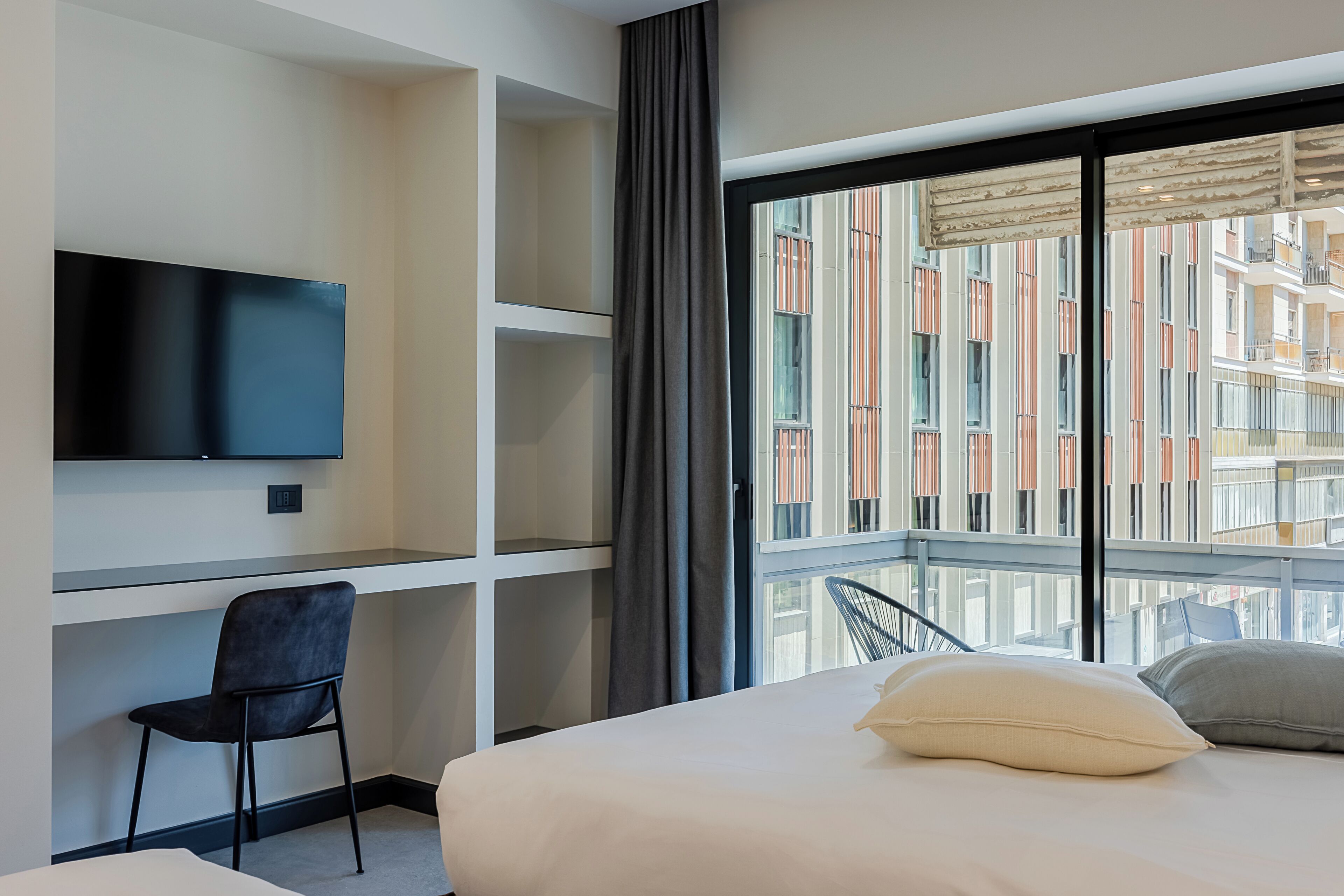 A hotel room with a bed, a desk, a chair, a TV, and a large window with a view of buildings.