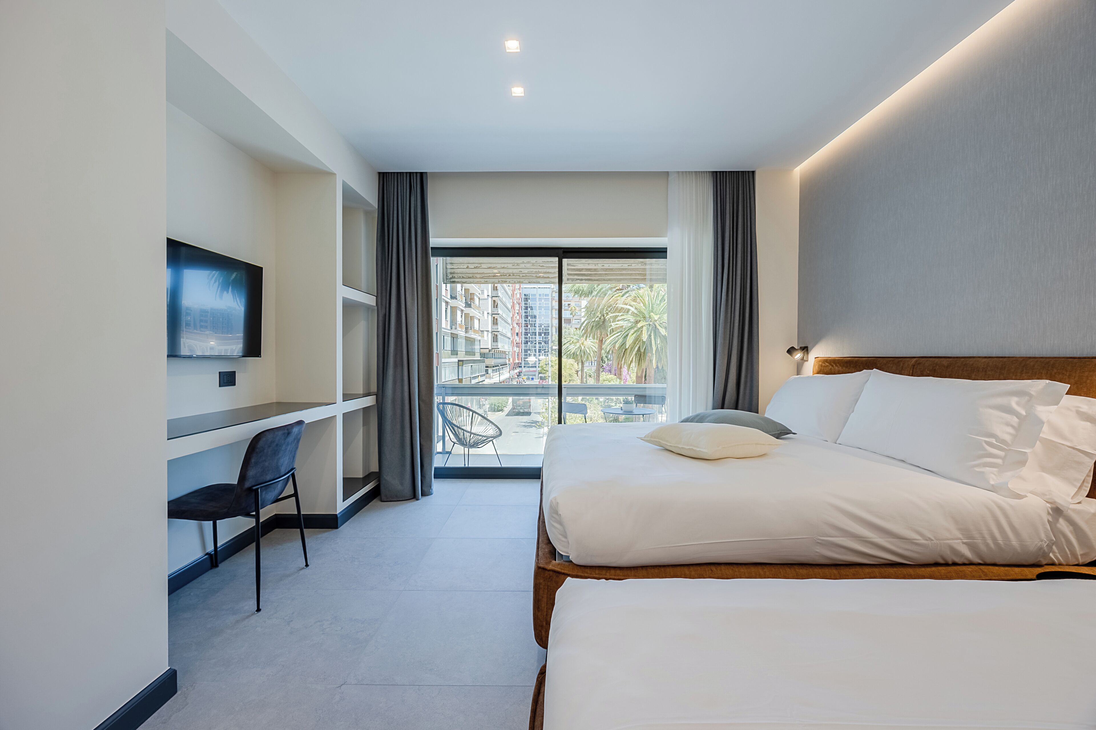 A modern hotel room with a bed, a desk, a chair, a TV, and a balcony with a view of buildings.