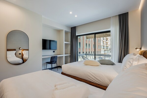 A hotel room with two beds, a desk, a chair, a TV, and a large window with a view of buildings.