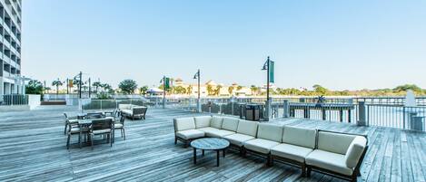 A wooden deck with outdoor seating, including a sofa and a round table, overlooking a beach area.
