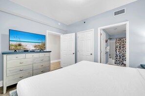 Une chambre à coucher comprenant un lit, une commode, un téléviseur diffusant une scène de plage, et deux portes donnant sur d’autres pièces.