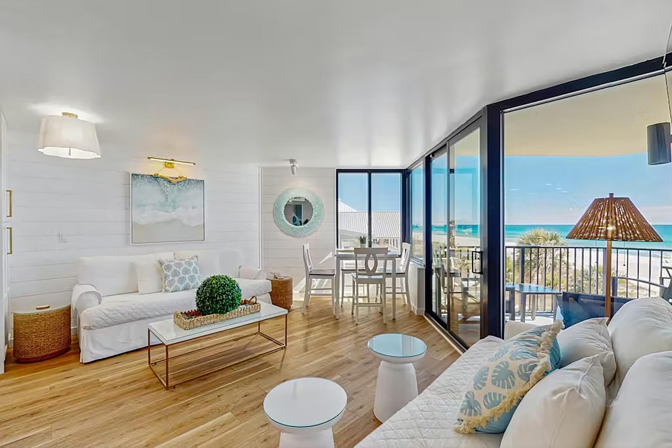 Ein modernes Wohnzimmer mit einem weißen Sofa, einem Couchtisch und einem Essbereich mit einem Tisch mit Glasplatte. Ein großes Fenster bietet Blick auf den Strand.