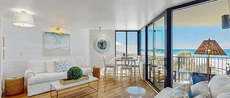 Una sala de estar moderna con un sofá blanco, una mesa de centro y un comedor con una mesa de vidrio. Se ve una ventana grande que ofrece vistas a la playa.
