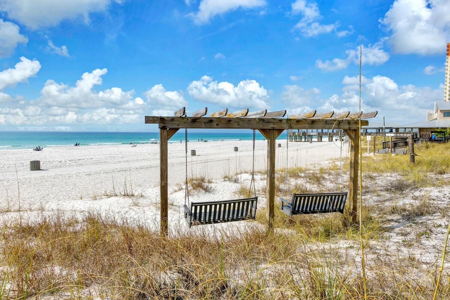 Une plage avec une structure en bois comprenant des balançoires et des bancs, une plage de sable, et une jetée en arrière-plan.