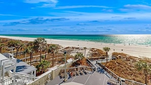 En ejendom ved stranden med en træpromenade, der fører til et poolområde, med udsigt over en sandstrand og havet.