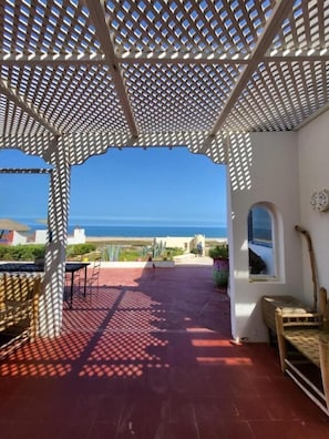 Un espacio al aire libre cubierto con vistas al mar, un patio con mesa y sillas, y un banco de madera.