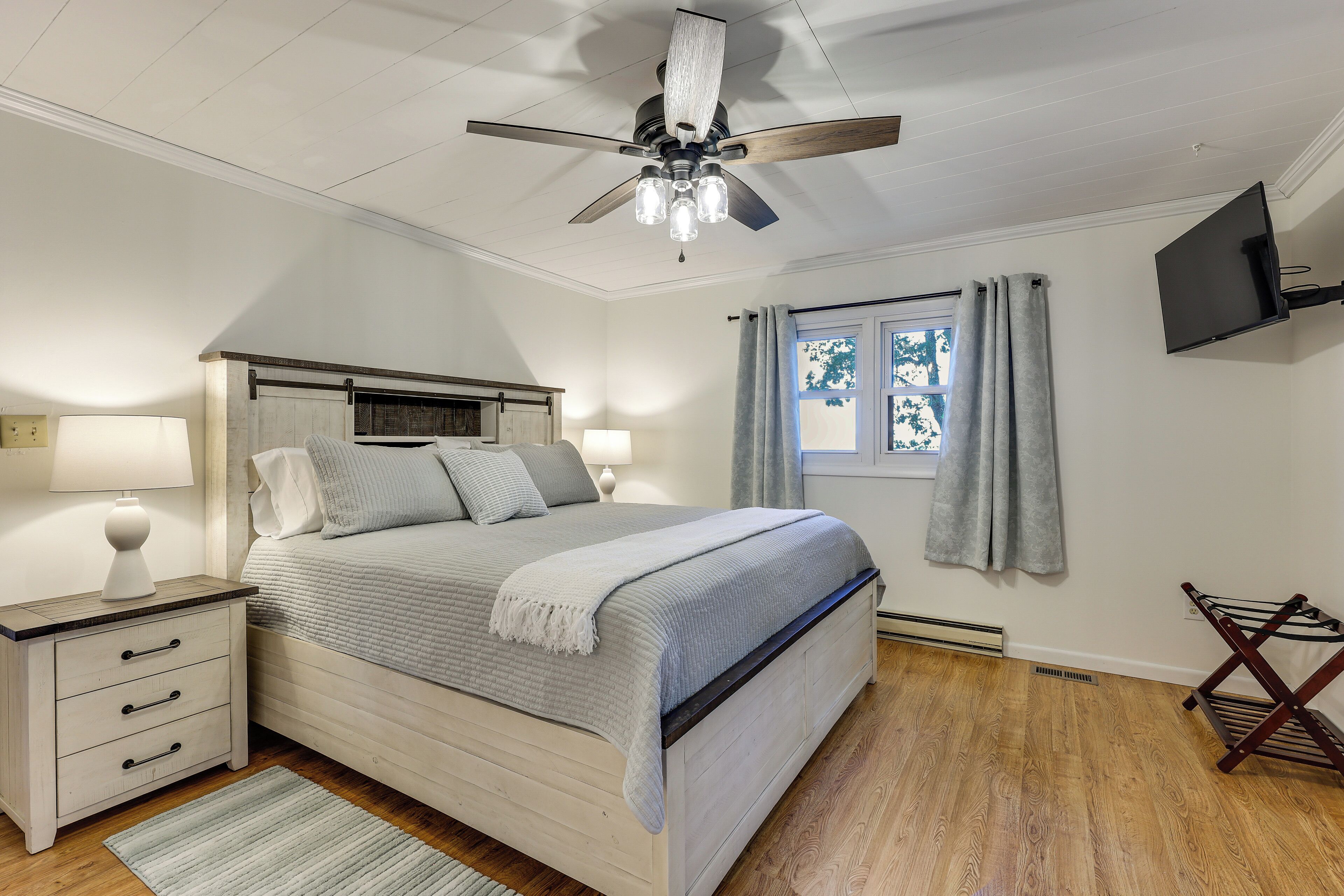 A bedroom with a bed, nightstands, a ceiling fan, a window with curtains, and a flat-screen TV mounted on the wall.