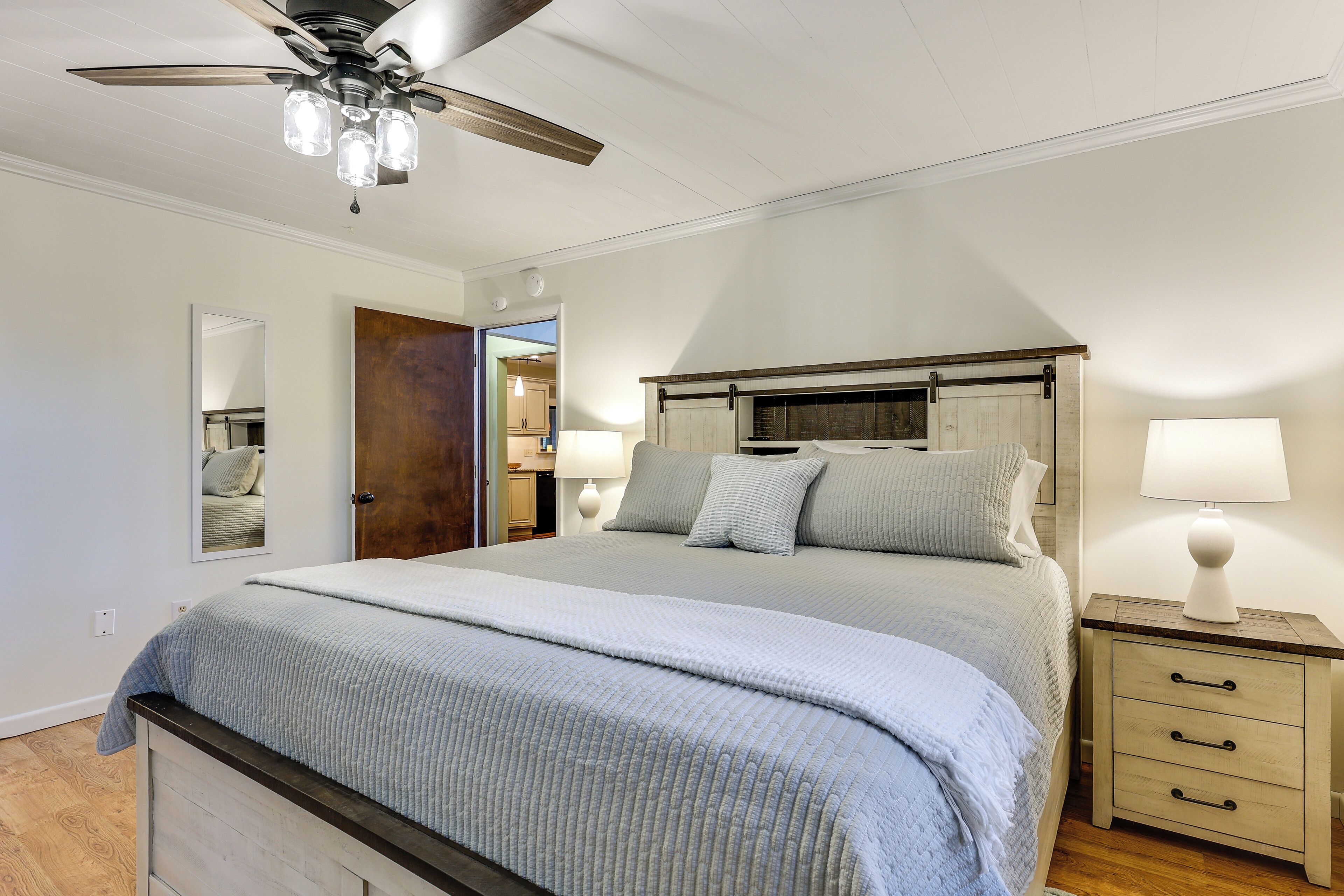 Un dormitorio con una cama, mesitas de noche, ventilador de techo y una puerta que da a un baño.