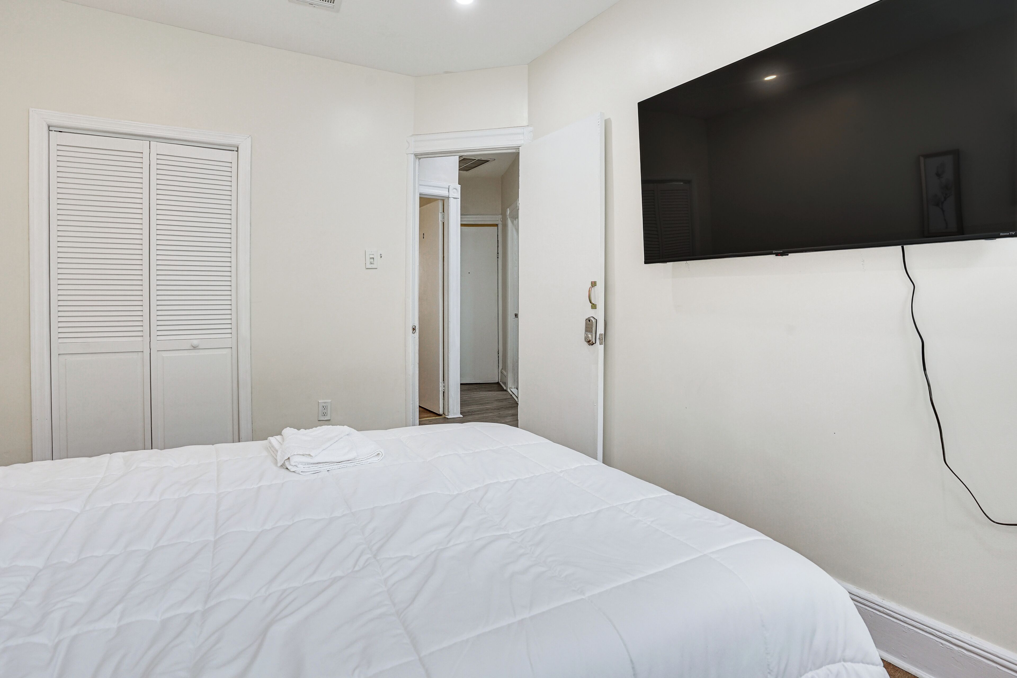 Un dormitorio con cama, televisor montado en la pared, una puerta y un pasillo.