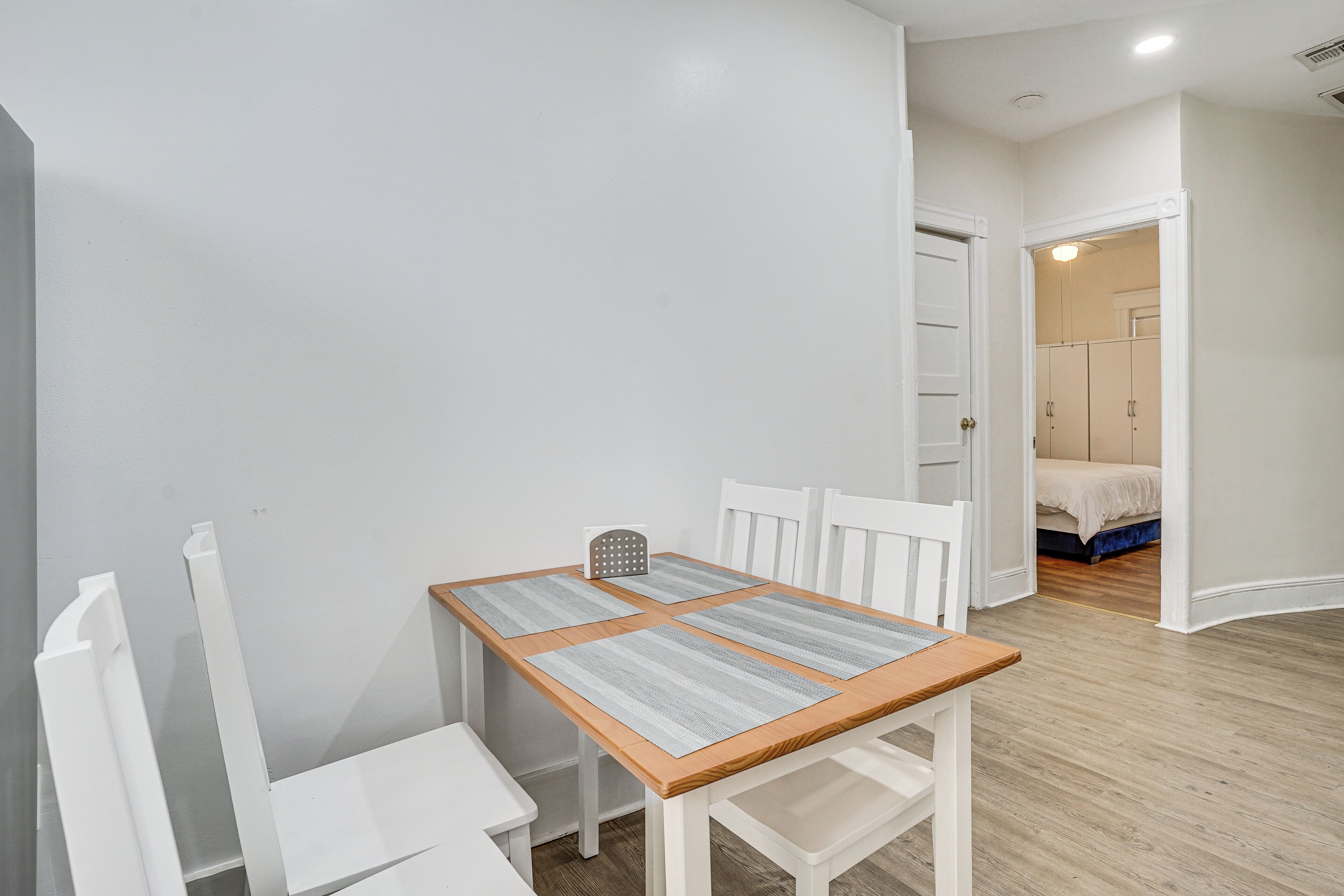 Quarto com mesa de madeira, cadeiras brancas, uma mesinha pequena branca e uma cama com colchão azul ao fundo.
