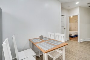 Una habitación con una mesa de madera, sillas blancas, una mesita pequeña y una cama con colchón azul al fondo.