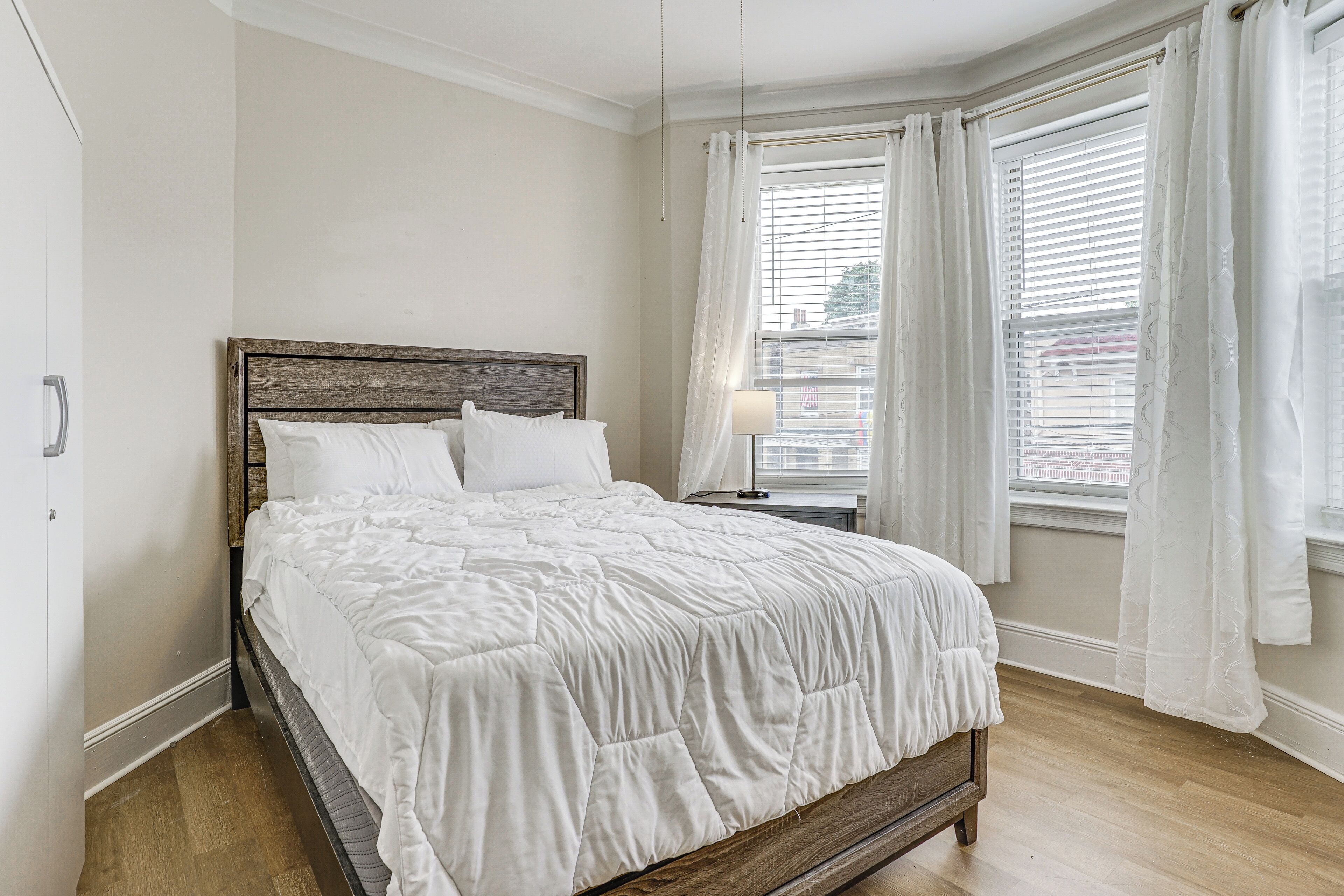Une chambre à coucher avec un lit en bois, du linge de lit blanc, une table de chevet et une fenêtre avec des stores.