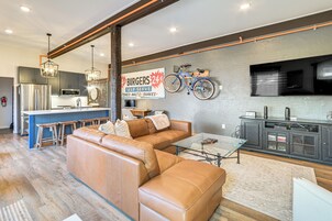 Una sala moderna con un sofá de cuero, una mesa de centro de vidrio y una zona de cocina con refrigerador, microondas y un letrero que anuncia hamburguesas.