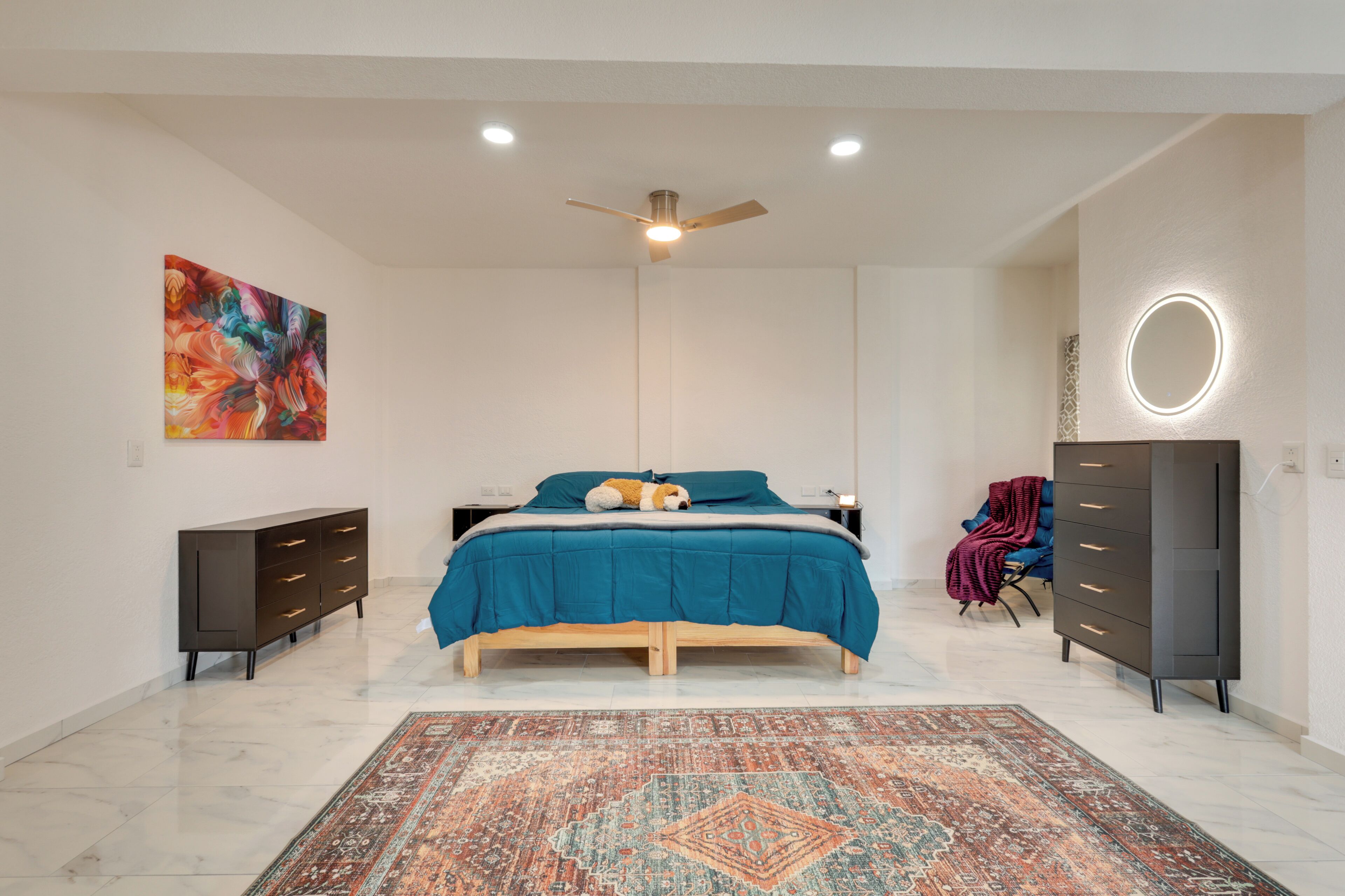 Un dormitorio con cama, cómoda, silla, alfombra, ventilador de techo y un cuadro.