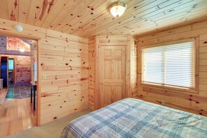 Une pièce de cabane en bois comprenant un lit, une porte, une fenêtre avec des stores et un luminaire au plafond.