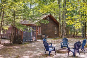 Eine rustikale Holzhütte mit Veranda, umgeben von Bäumen und einem Feuerkorb mit Sitzgelegenheiten.