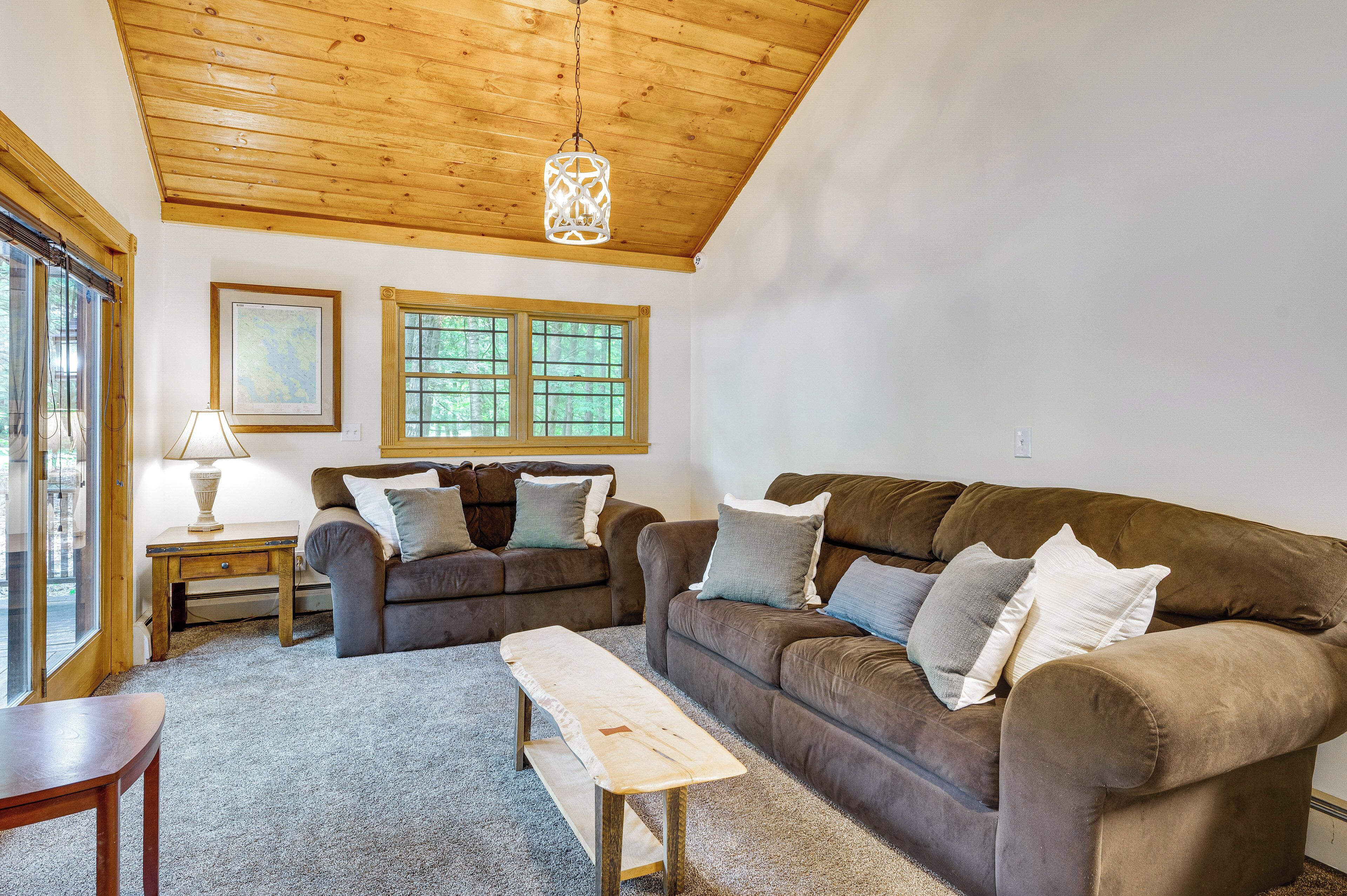 Ein gemütliches Wohnzimmer mit einem braunen Sofa, einem Holztisch und einer Lampe. Zwei Fenster bieten Blick auf Bäume.