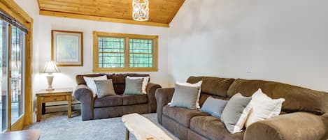 Ein gemütliches Wohnzimmer mit einem braunen Sofa, einem Holztisch und einer Lampe. Zwei Fenster bieten Blick auf Bäume.
