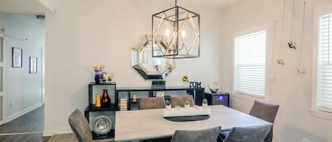Uma sala de jantar com uma mesa retangular, seis cadeiras, uma luminária pendente hexagonal e um aparador preto com objetos decorativos.