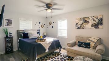 A bedroom with a bed, a ceiling fan, a window, a chair, and a wall with a painting.