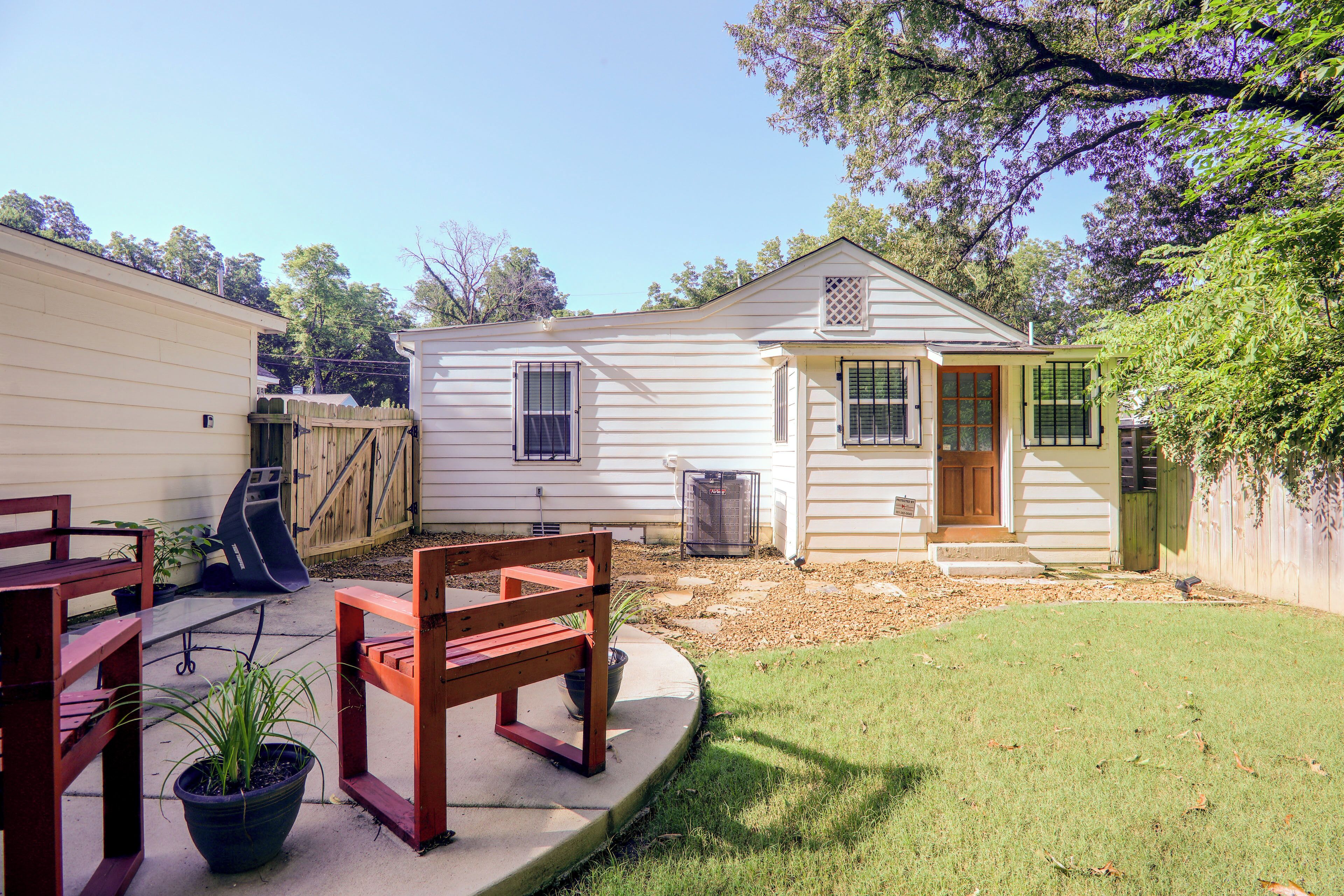 A residential backyard with a wooden deck, a red bench, a potted plant, and a small white house with a front door and windows.