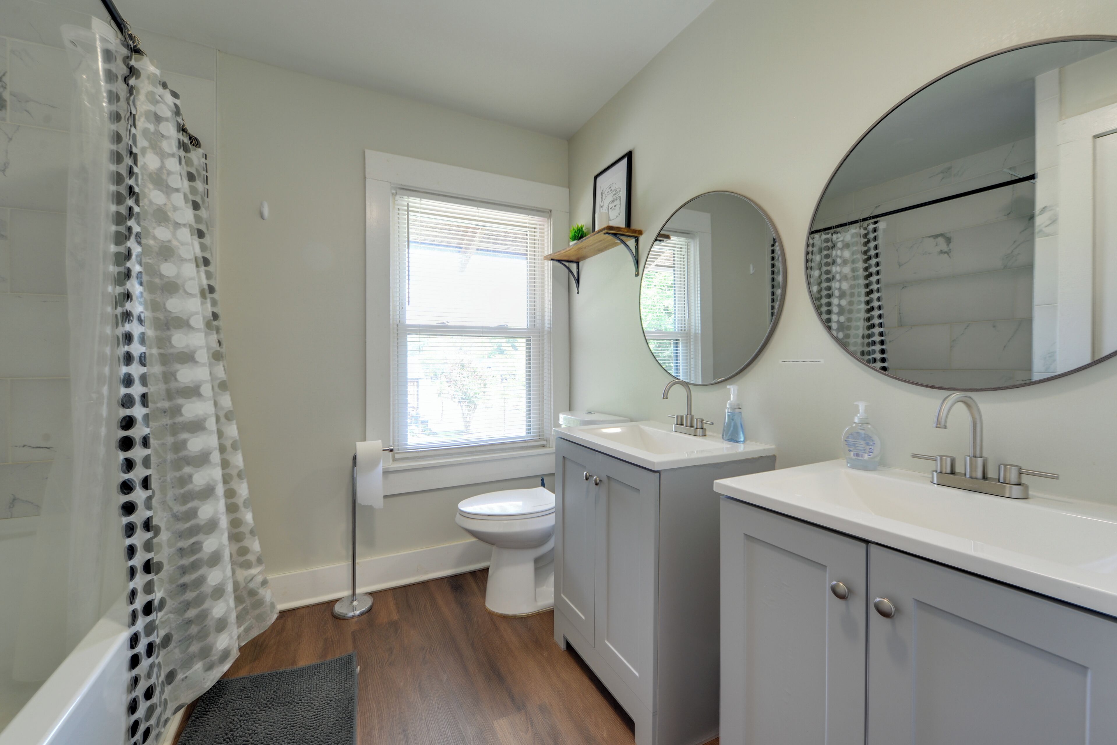 Une salle de bain avec des toilettes, un lavabo et un miroir. Il y a une fenêtre avec des stores, un rideau de douche et une petite étagère avec une plante.