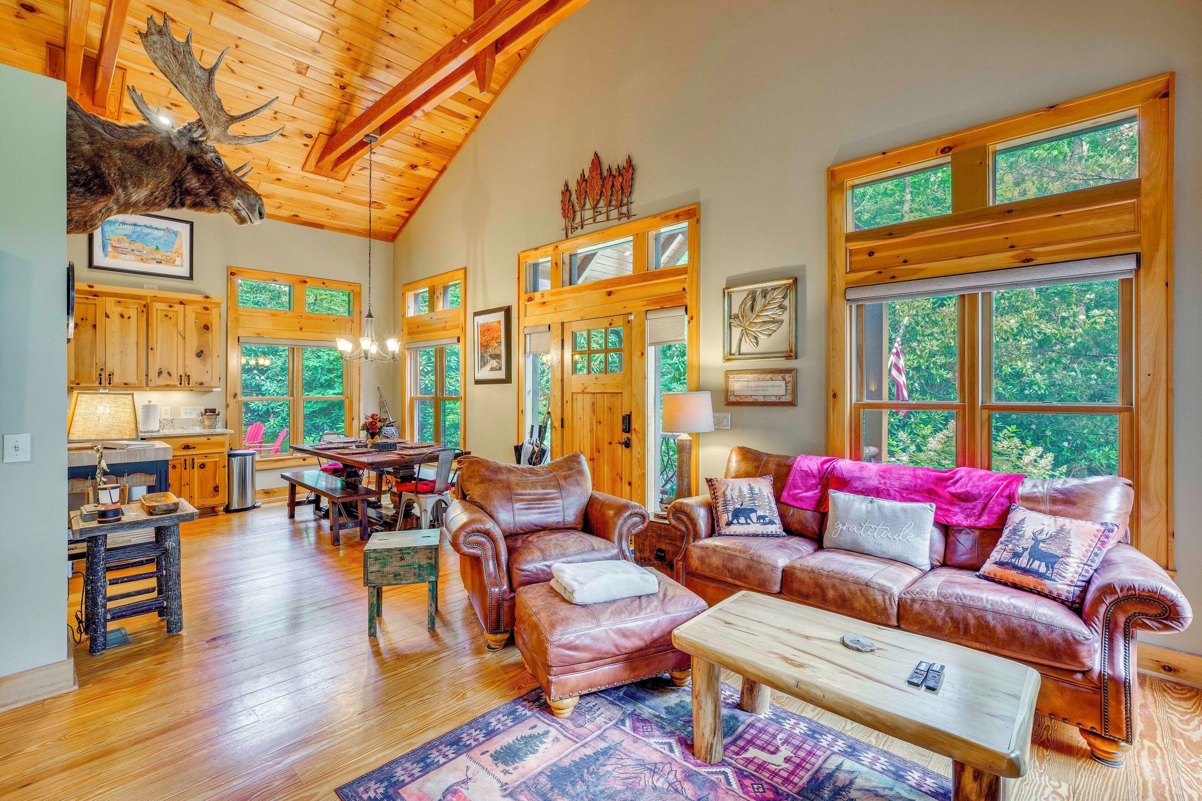 A spacious living room with a high ceiling, wooden beams, large windows, a fireplace, and a cozy seating area with leather furniture.