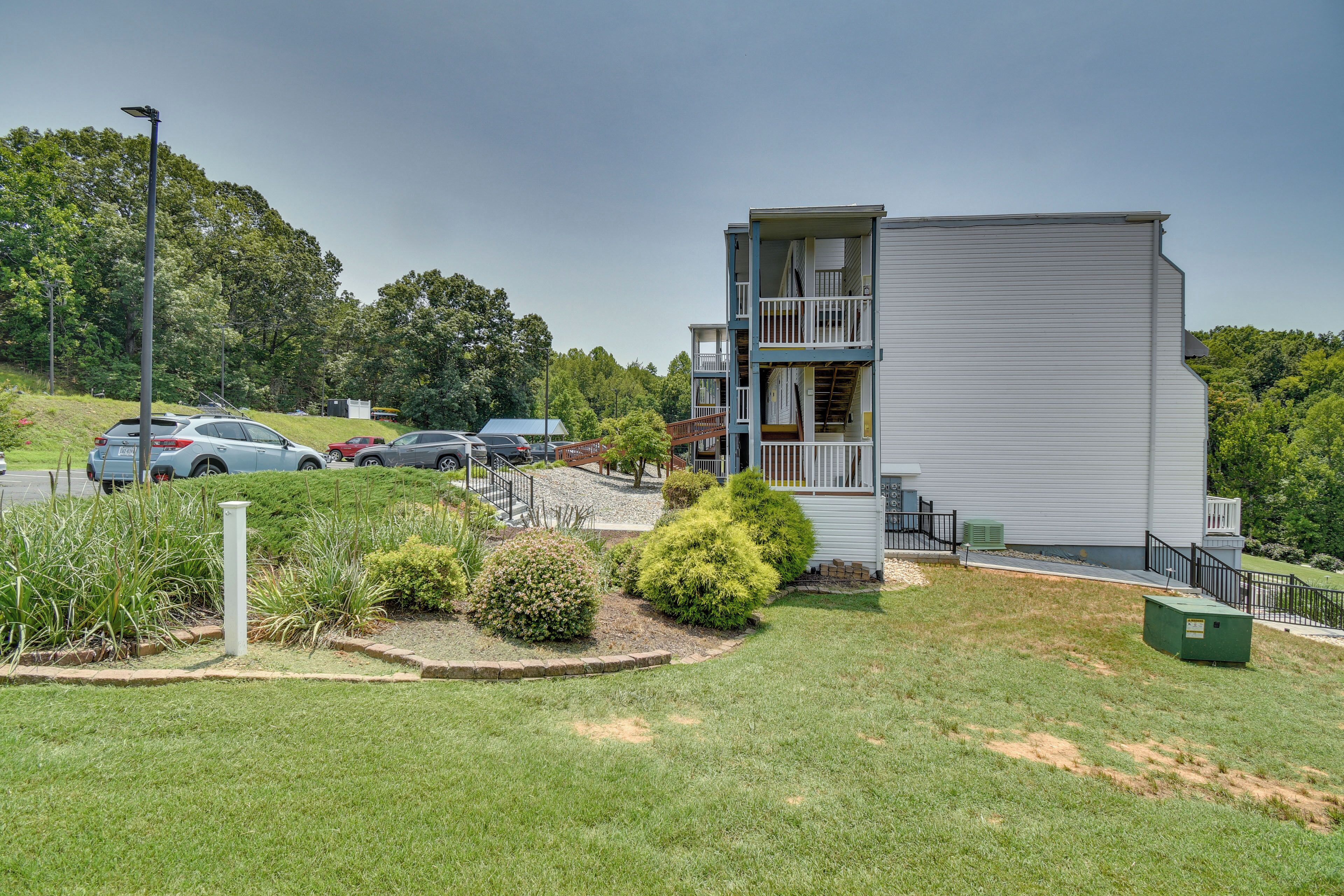 A multi-story building with a flat roof, a parking area with several cars, and a grassy area with shrubs.