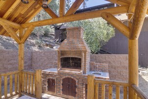 Cocina exterior de ladrillo con chimenea, terraza de madera y un muro de piedra al fondo.
