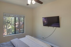 Une chambre à coucher avec un lit, un ventilateur de plafond, un téléviseur fixé au mur et une fenêtre avec des stores.