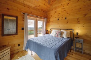 A bedroom with a wooden ceiling, a bed with a blue bedspread, a nightstand, and a window with a view of greenery. - 66-acre Group Escape! Gateway to Grandfather Mtn (Elk Park)