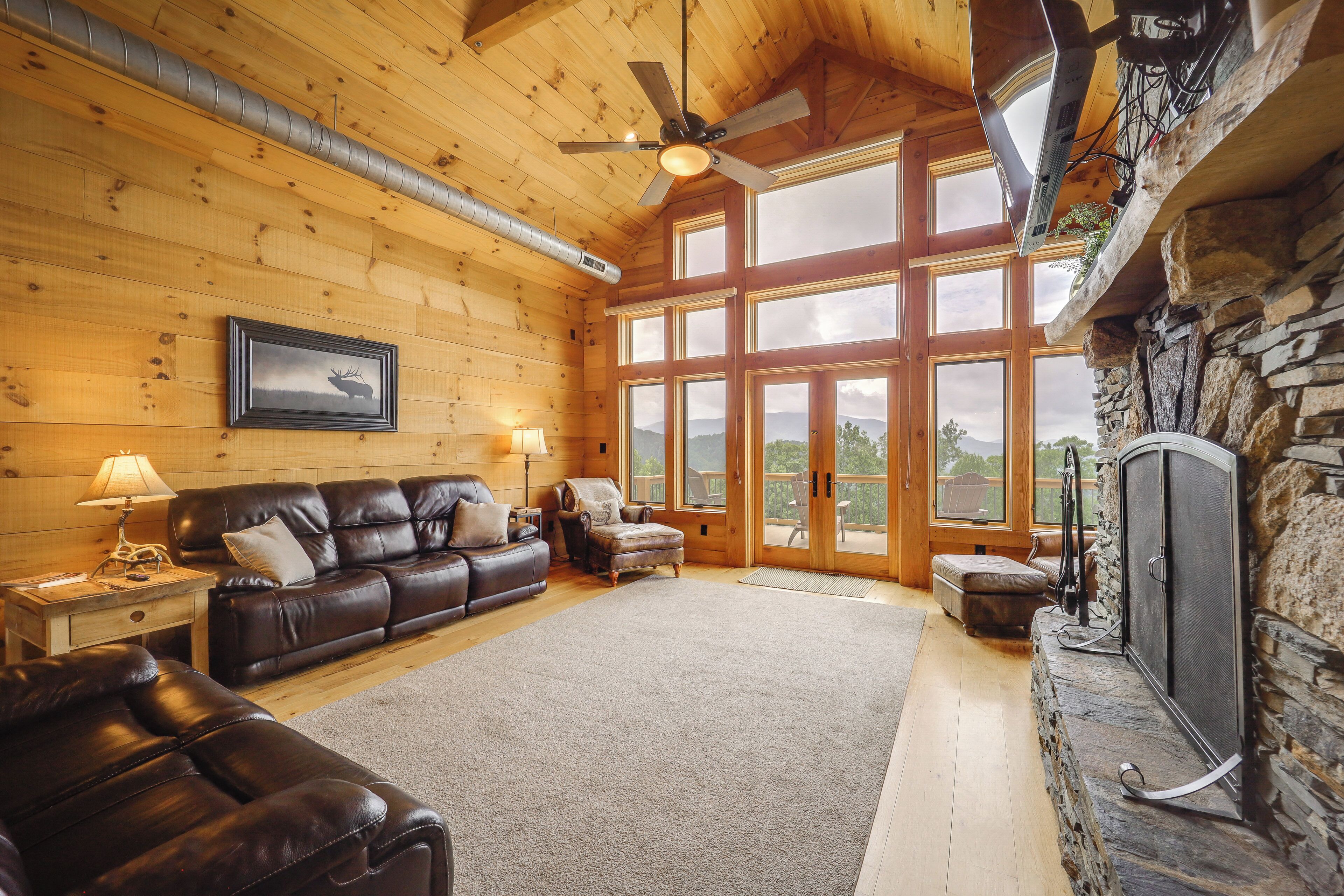 A log cabin interior with a leather sofa, a fireplace, and a large window overlooking a mountainous landscape.