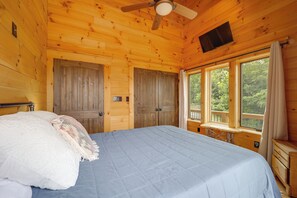 Quarto de cabana de madeira com uma cama, um ventilador de teto, uma janela com cortinas e uma televisão na parede.