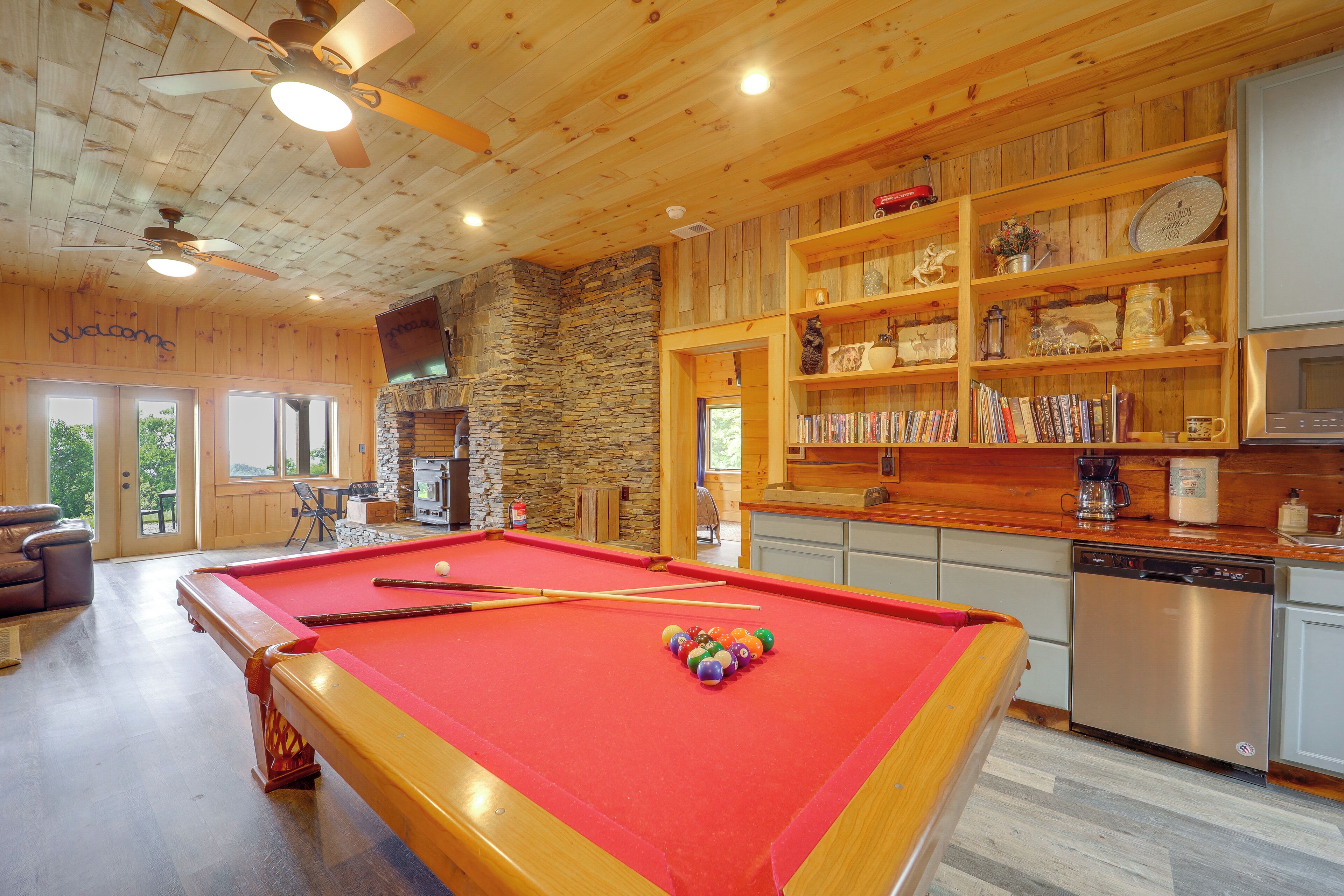 A pool table with pool cues and balls, a stone fireplace, wooden shelves with decorative items, and a kitchen area with a dishwasher.