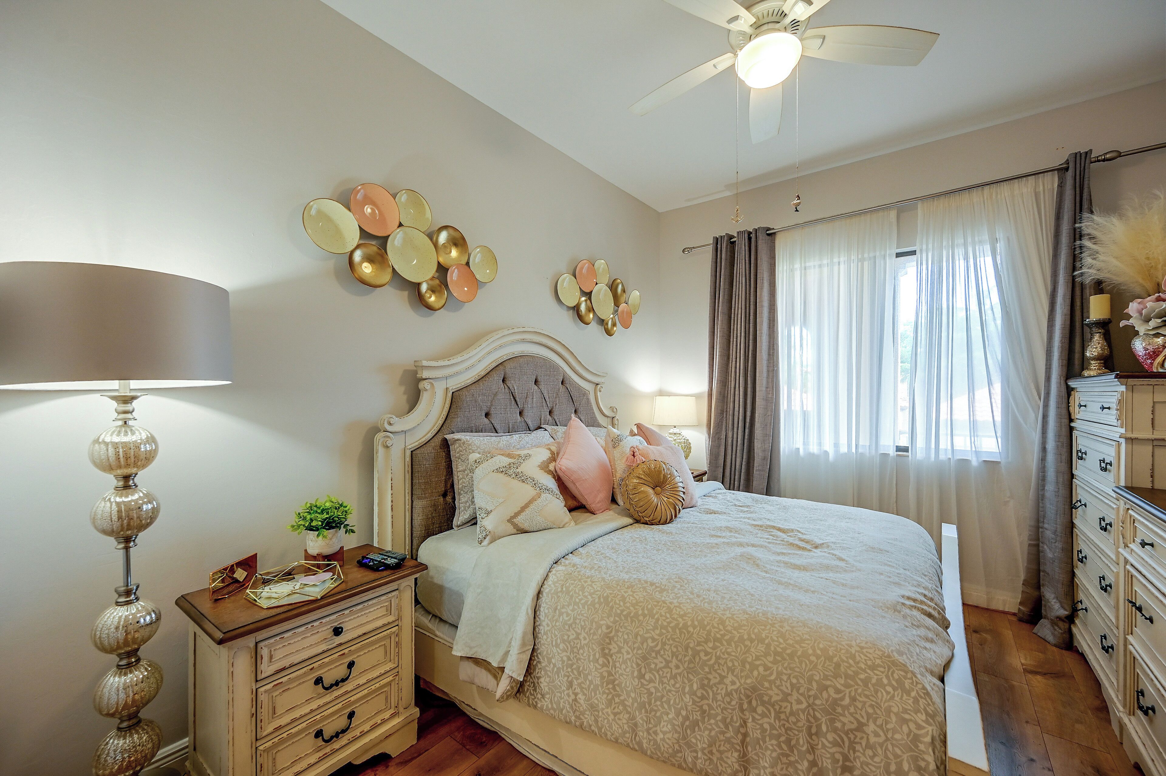 Une chambre à coucher comprenant un lit, une table de chevet, une lampe, des œuvres d’art décoratives au mur, des rideaux et une commode.