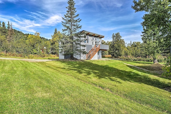 A modern two-story house with a wooden staircase, surrounded by a well-maintained lawn and tall trees.