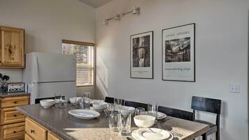 A kitchen with granite countertops, wooden cabinets, and a dining area with a table set for four. There are framed pictures on the wall and a window with blinds.