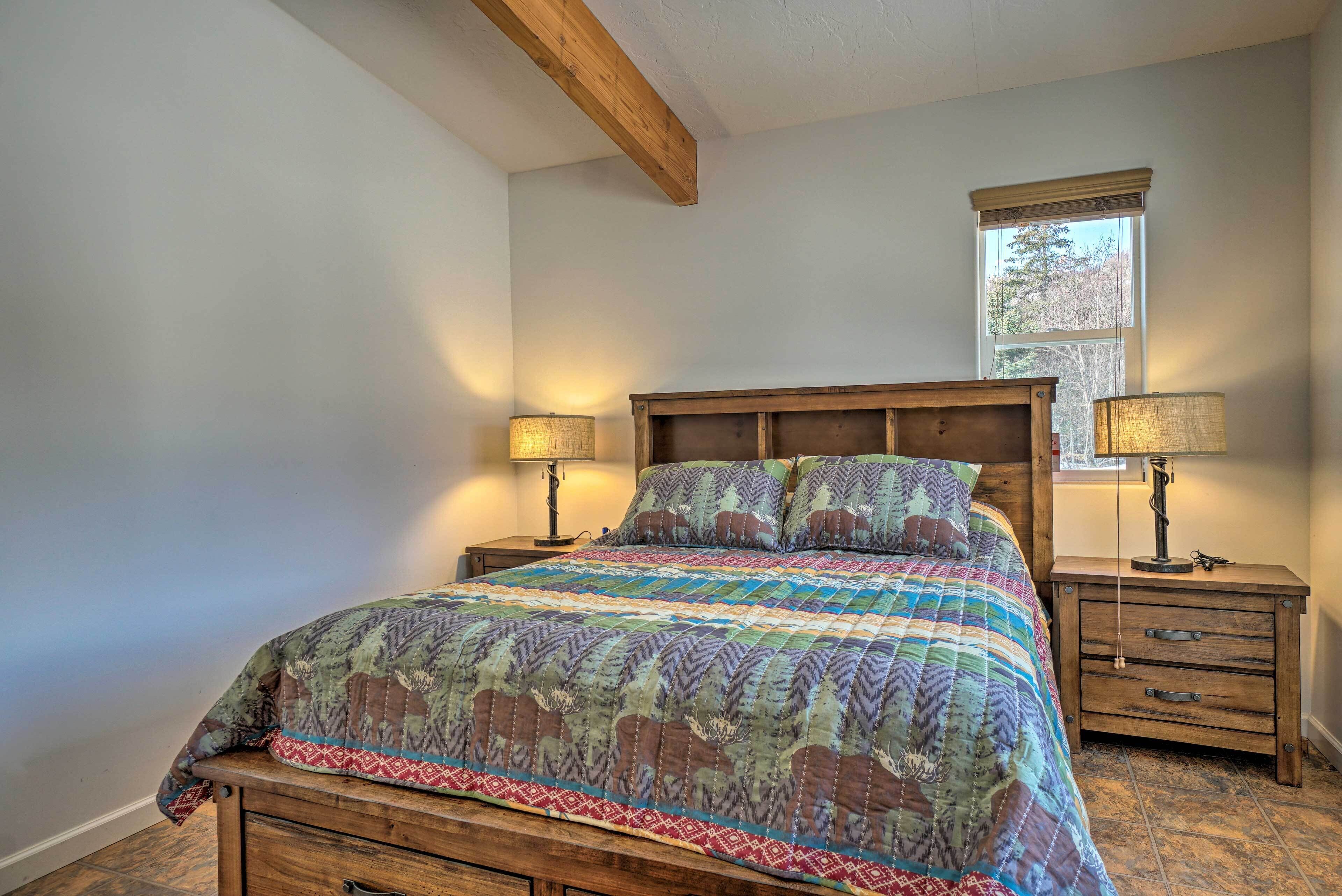 A bedroom with a large bed, wooden headboard, two bedside tables with lamps, a window with blinds, and a wooden dresser.