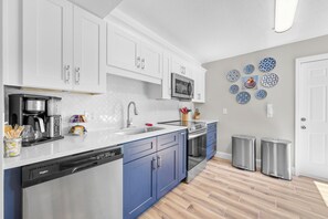 A modern kitchen with white cabinets, a stainless steel dishwasher, a microwave, and a stove. There are two trash bins and a set of blue cabinets. - 13 Mi to Beach! Family-friendly Palm Bay Retreat (Palm Bay)