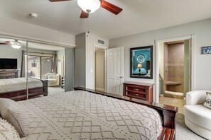 Ein Schlafzimmer mit einem großen Bett, einem Deckenventilator, einem Schminktisch, einem Sessel und einer Tür zu einem anderen Raum.