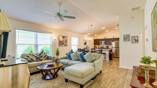 A spacious living room with a ceiling fan, a dining area with a kitchen in the background, and a large window providing natural light. - Community Pools, Golf & More! Home in The Villages (Wildwood)