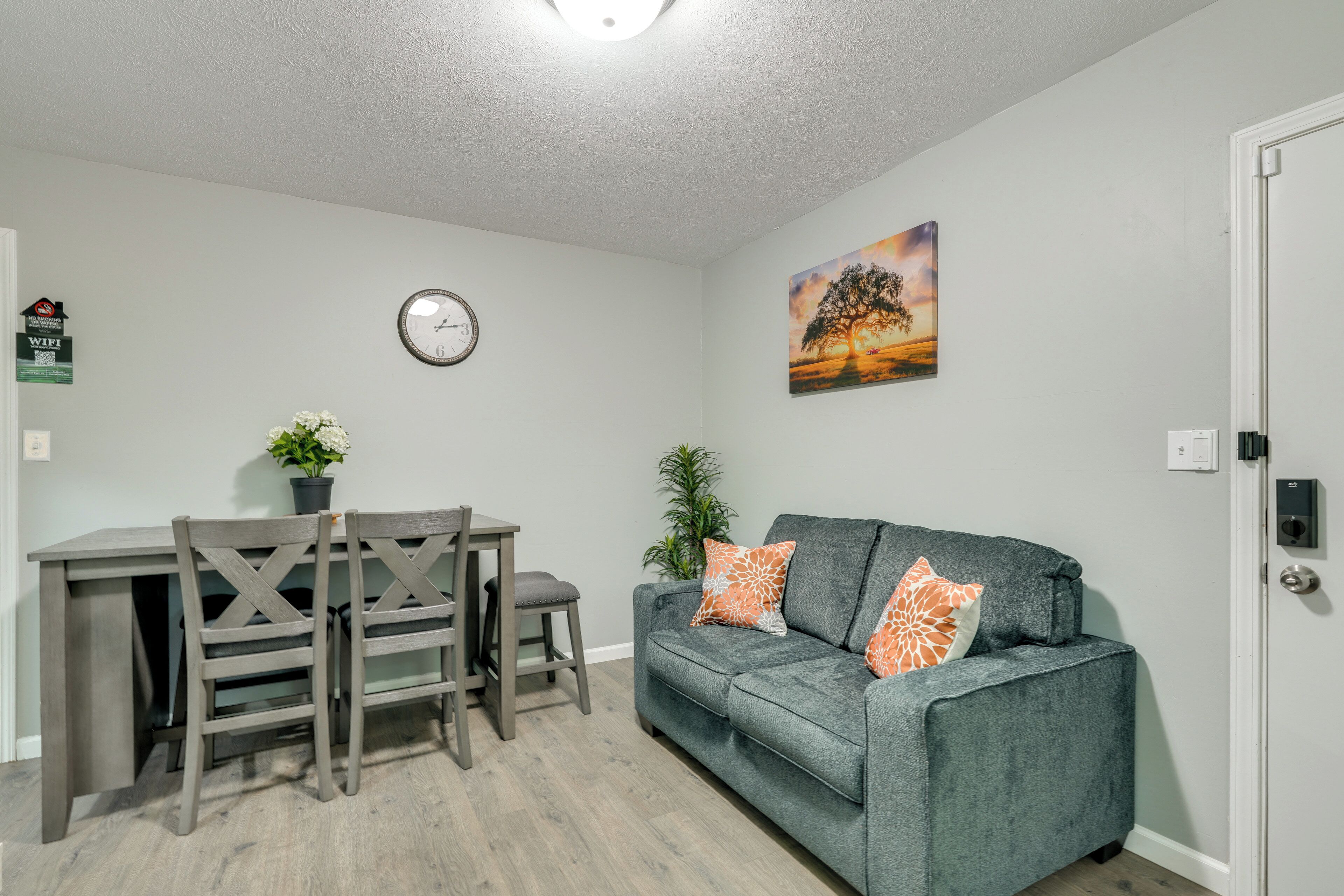 A living room with a grey sofa, a dining table with chairs, a wall clock, and a painting.