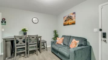 A living room with a grey sofa, a dining table with chairs, a wall clock, and a painting.