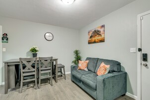 A living room with a grey sofa, a dining table with chairs, a wall clock, and a painting.