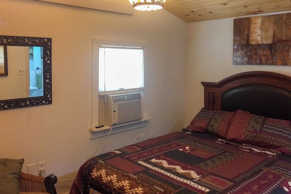 A bedroom with a bed, a window with an air conditioning unit, a mirror, and a wall-mounted artwork.